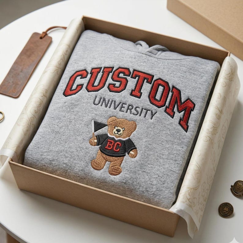 A Gildan Sport Grey hoodie neatly folded inside a luxury kraft gift box with premium tissue paper. The custom university embroidery features red "CUSTOM" lettering and an embroidered bear mascot. Ideal for personalized graduation gifts or alumni homecoming presents, this high-end presentation highlights the quality of custom-made collegiate outerwear.
