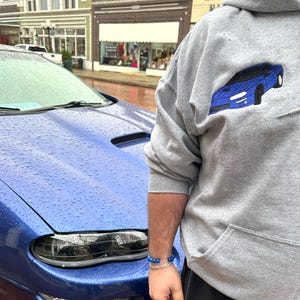 Sport grey embroidered hoodie featuring a detailed blue sports car design, shown next to the actual car. Personalized car sweatshirt for car lovers and custom automotive gift idea for men.