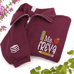 Custom embroidered maroon quarter-zip sweatshirt with “Ms. Freya” and cute apple, pencil, and ruler design, plus white book stack on sleeve — personalized teacher sweatshirt, perfect back-to-school outfit, thoughtful teacher appreciation gift, or cozy classroom wear.