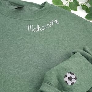 Custom embroidered heather dark green crewneck sweatshirt featuring white 'Mahanoy' text on the neckline and a black-and-white soccer ball design on the sleeve. Perfect personalized sports team apparel, soccer fan gift, or school spirit wear. Cozy unisex sweatshirt ideal for game days, casual outfits, and custom embroidery lovers.