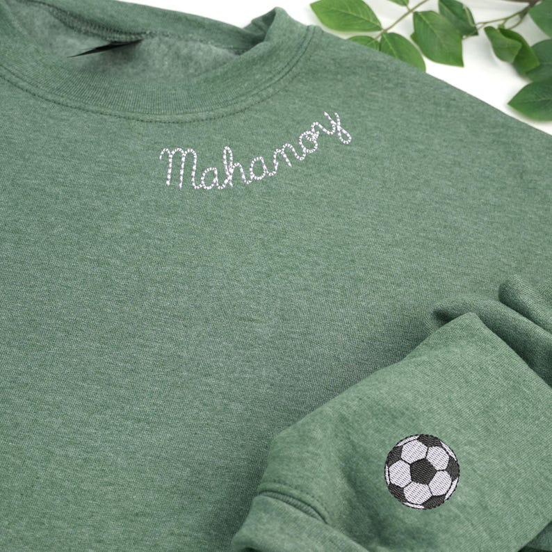 Custom embroidered heather dark green crewneck sweatshirt featuring white 'Mahanoy' text on the neckline and a black-and-white soccer ball design on the sleeve. Perfect personalized sports team apparel, soccer fan gift, or school spirit wear. Cozy unisex sweatshirt ideal for game days, casual outfits, and custom embroidery lovers.