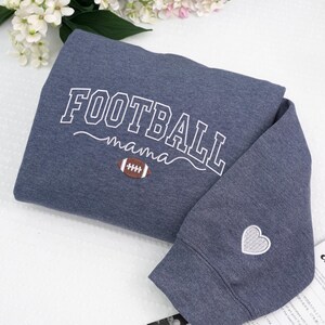 Custom embroidered Football Mama sweatshirt in Heather Dark Navy, featuring varsity lettering with football design on chest and white heart embroidery on sleeve. Soft cotton-poly blend for cozy everyday wear. Perfect game day outfit, thoughtful gift for football moms, or team spirit apparel. Handmade embroidery adds a premium, one-of-a-kind touch. Available in multiple sizes and colors.