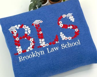 May include: A blue sweatshirt featuring embroidered letters B, L, and S formed by red leaves and white flowers, with small graduation caps atop each letter. The text Brooklyn Law School is embroidered in white below the large letters.