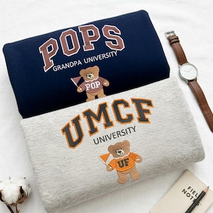 Two folded Gildan sweatshirts in Navy and Sport Grey. The navy sweatshirt features "POPS Grandpa University" embroidery, while the grey features "UMCF University." Both include a custom mascot bear with a matching sweater. These personalized grandpa gifts and collegiate-style sweatshirts are essential for family-themed apparel and custom school spirit collections.