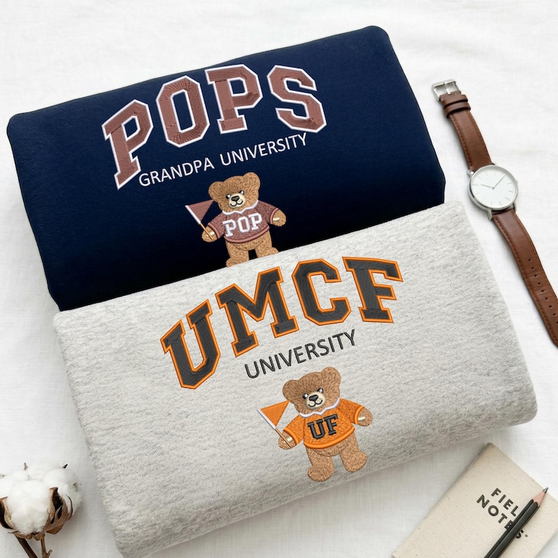 Two folded Gildan sweatshirts in Navy and Sport Grey. The navy sweatshirt features "POPS Grandpa University" embroidery, while the grey features "UMCF University." Both include a custom mascot bear with a matching sweater. These personalized grandpa gifts and collegiate-style sweatshirts are essential for family-themed apparel and custom school spirit collections.