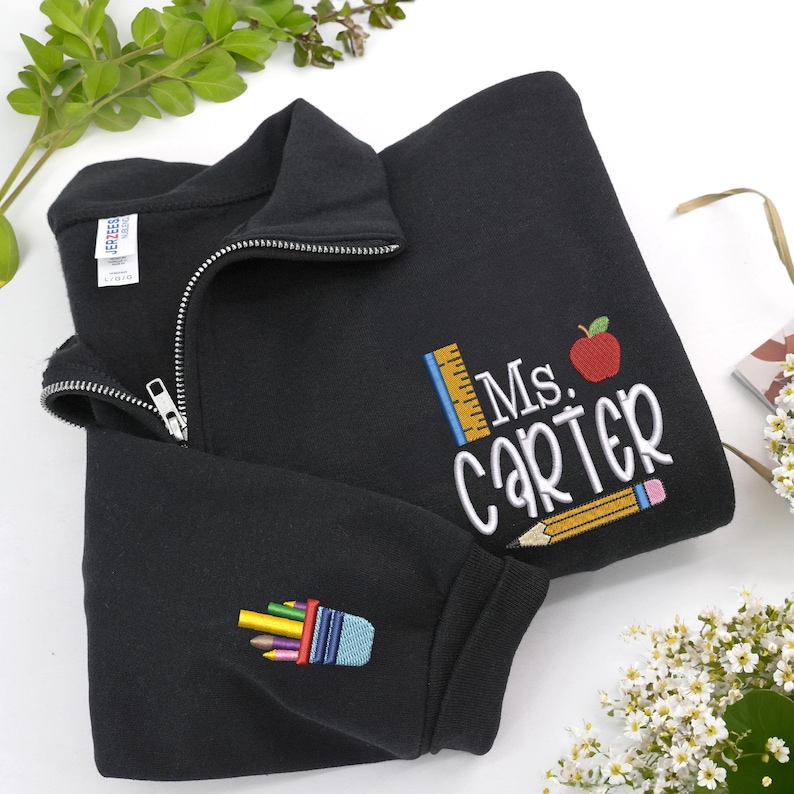 Custom embroidered black quarter-zip sweatshirt with “Ms. Carter” and cute apple, pencil, and ruler design, plus colorful pencil case sleeve embroidery — personalized teacher sweatshirt, perfect back-to-school outfit, thoughtful teacher appreciation gift, or cozy classroom wear.