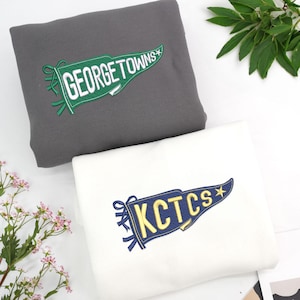 Peut inclure: Deux sweat-shirts pliés, un gris et un blanc. Le sweat-shirt gris porte une bannière brodée verte et blanche avec l'inscription "GEORGETOWNS". Le sweat-shirt blanc porte une bannière brodée bleue et jaune avec l'inscription "KCTCS".