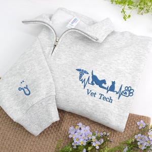 Op de afbeelding: Een lichtgrijs Vet Tech sweatshirt met een kraag met een kwartrits. De sweatshirt heeft een blauwe stethoscoop op de linkermouw en een blauw geborduurd ontwerp met de woorden "Vet Tech" en een hond, kat en pootafdruk.
