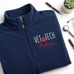 May include: A navy blue fleece jacket with a full zipper, embroidered with "VET TECH" in white, a red paw print, and the name "Rishima" in red script. A stethoscope, pen, and small plant are in the background.