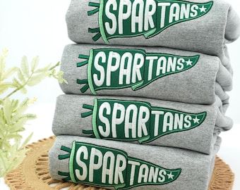 Könnte beinhalten: Ein Stapel von vier grauen Sweatshirts. Jedes Sweatshirt hat ein grün-weißes Wimpeldesign mit dem gestickten Wort "SPARTANS". Die Sweatshirts sind ordentlich gefaltet und übereinander gestapelt.