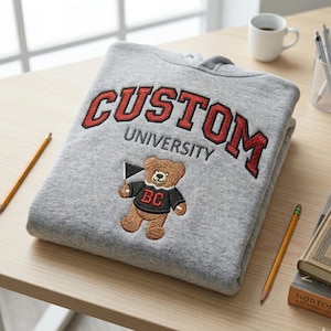 May include: A folded gray hoodie with the embroidered text "CUSTOM UNIVERSITY" in red and black. A brown teddy bear with a black shirt and "BC" on it is embroidered below the text. A pencil and a coffee cup are on the table.