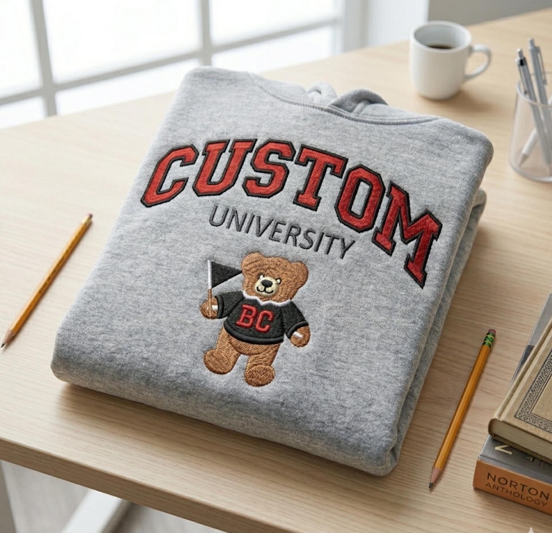 A Gildan Sport Grey heavy blend hoodie featuring red custom varsity letter embroidery and a bear mascot. The hoodie is folded on a desk with pencils and a "Norton Anthology" book. This personalized school spirit apparel is perfect for back-to-school gift guides and custom orders, providing a high-quality, timeless university campus aesthetic.