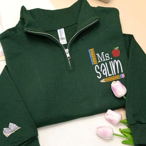 Custom embroidered forest green quarter-zip sweatshirt with “Ms. Salim” and cute apple, pencil, and ruler design, plus open book sleeve embroidery — personalized teacher sweatshirt, perfect back-to-school outfit, thoughtful teacher appreciation gift, or cozy classroom wear.