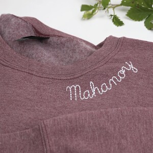 Heather dark maroon Gildan sweatshirt with custom embroidered text 'Mahanoy' in white cursive on the collar. Soft, cozy unisex crewneck perfect for personalized school spirit wear, sports team apparel, or thoughtful custom gifts. Ideal for students, alumni, and fans looking for unique embroidered clothing for game day, casual wear, or special occasions