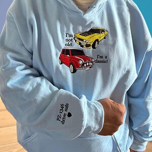 Light blue embroidered hoodie featuring two classic car designs with the quote “I’m not old, I’m a classic!” and custom sleeve text “PZ-4346 drive safe.” Perfect personalized car lover gift for vintage car enthusiasts.