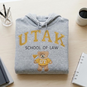 Overhead flat lay of a Gildan Sport Grey hoodie with "UTAK SCHOOL OF LAW" custom mascot embroidery. The gold and grey thread colors create a premium collegiate look. A must-have visual for custom apparel shops, this image targets keywords like personalized university merch, law school spirit wear, and custom embroidered bear hoodies.