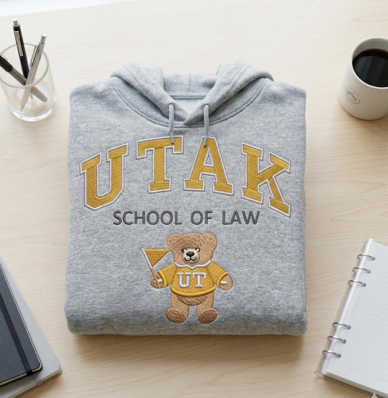 Overhead flat lay of a Gildan Sport Grey hoodie with "UTAK SCHOOL OF LAW" custom mascot embroidery. The gold and grey thread colors create a premium collegiate look. A must-have visual for custom apparel shops, this image targets keywords like personalized university merch, law school spirit wear, and custom embroidered bear hoodies.