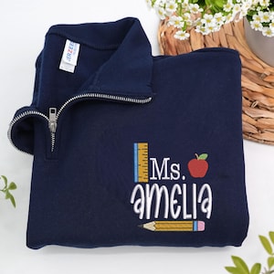 Custom embroidered navy quarter-zip sweatshirt with “Ms. Amelia” and cute apple, pencil, and ruler design — personalized teacher sweatshirt, ideal for back-to-school, thoughtful teacher appreciation gift, or cozy everyday classroom wear.