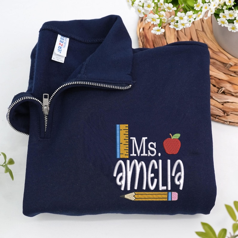 Custom embroidered navy quarter-zip sweatshirt with “Ms. Amelia” and cute apple, pencil, and ruler design — personalized teacher sweatshirt, ideal for back-to-school, thoughtful teacher appreciation gift, or cozy everyday classroom wear.