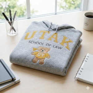 A Gildan Sport Grey heavy blend hoodie folded on a light wood desk next to a notebook and coffee mug. The custom law school embroidery features "UTAK" in gold and a stitched bear mascot. This aesthetic university desk setup photography is perfect for marketing personalized student gifts and custom collegiate apparel with high-detail embroidery.