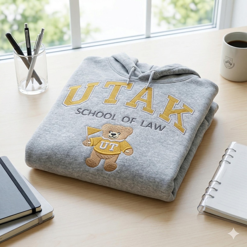A Gildan Sport Grey heavy blend hoodie folded on a light wood desk next to a notebook and coffee mug. The custom law school embroidery features "UTAK" in gold and a stitched bear mascot. This aesthetic university desk setup photography is perfect for marketing personalized student gifts and custom collegiate apparel with high-detail embroidery.