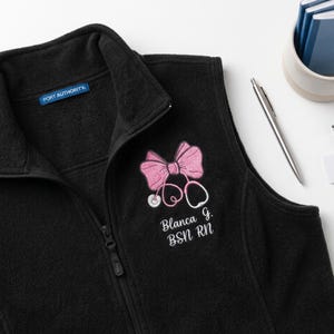 May include: A black fleece vest with a full-length zipper and a Port Authority label. Embroidered on the chest is a pink bow combined with a pink stethoscope design, followed by the white embroidered text: Blanca G. BSN RN.