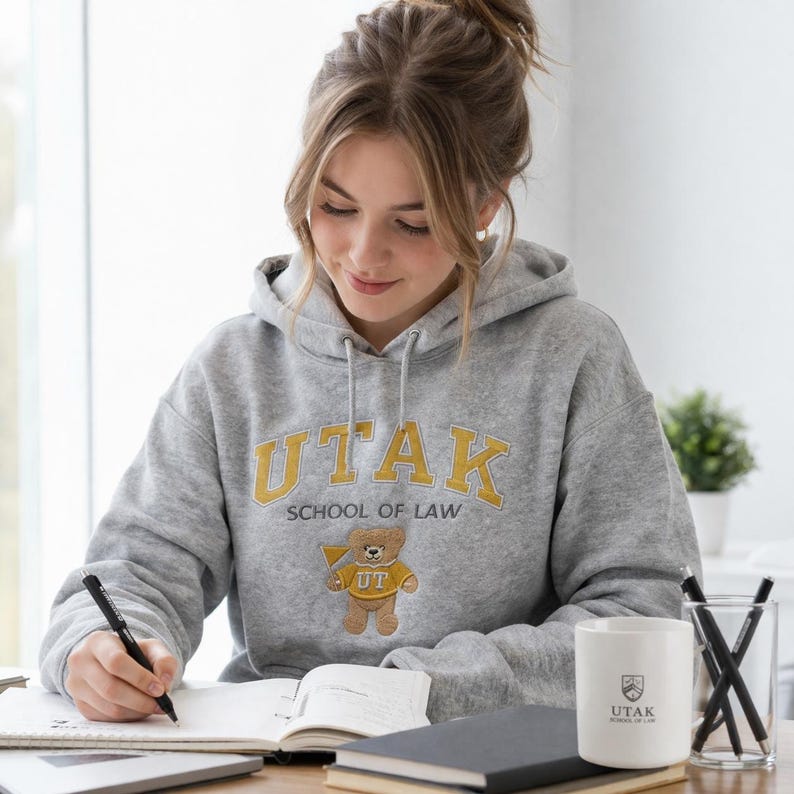 A female law student wearing a Gildan Sport Grey hoodie with "UTAK SCHOOL OF LAW" custom embroidery. The design includes a gold block letter logo and a charming bear mascot wearing a UT sweater. This personalized law school apparel is a top-trending graduation gift idea, offering a cozy and professional aesthetic for campus lifestyle and library study vibes.