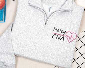 May include: A light gray quarter-zip sweatshirt with the name "Haley" and "CNA" embroidered in black, along with a pink heart-shaped EKG design. The sweatshirt has a silver zipper and a tag that reads "Jerzees".