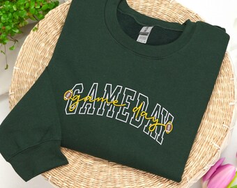 Può includere: Una felpa verde scuro con la scritta "GAMEDAY" in lettere bianche con dettagli gialli e una grafica di football americano. La felpa è esposta su un vassoio di vimini intrecciato con verde e fiori rosa.