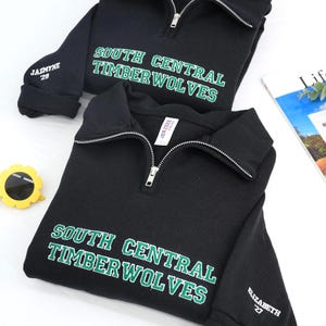 May include: Two black quarter-zip sweatshirts with the text "SOUTH CENTRAL TIMBERWOLVES" in green and white. One sweatshirt has "JAZMYNE '29" on the sleeve, the other has "ELIZABETH '27". A pair of yellow sunglasses and a magazine are in the background.