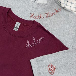 Set of two custom embroidered Gildan sweatshirts — sport grey with red 'Heath Hawks' text on the collar and football sleeve design with personalized name and number, and maroon with white 'Shalom' text on the collar. Cozy unisex crewnecks perfect for school spirit wear, sports teams, or personalized gifts. Ideal for game day, casual outfits, and custom apparel lovers.