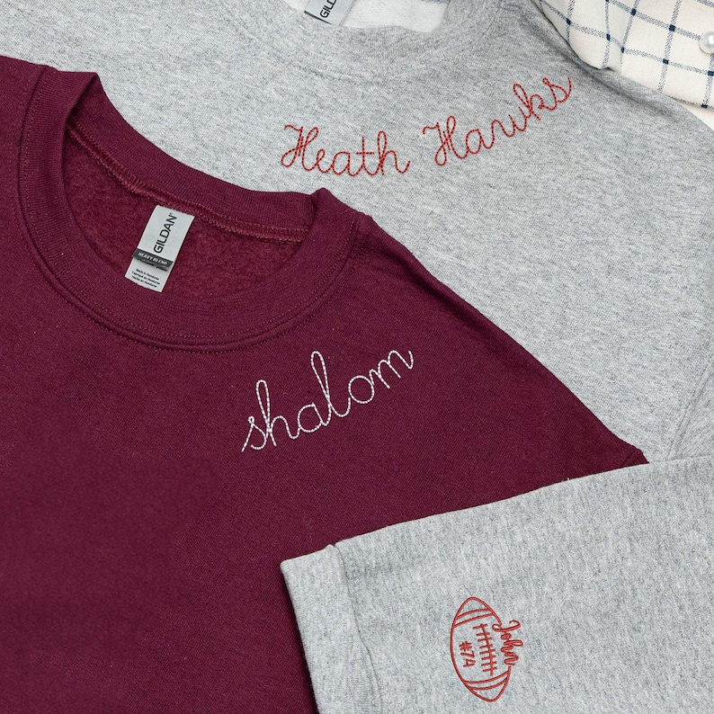 Set of two custom embroidered Gildan sweatshirts — sport grey with red 'Heath Hawks' text on the collar and football sleeve design with personalized name and number, and maroon with white 'Shalom' text on the collar. Cozy unisex crewnecks perfect for school spirit wear, sports teams, or personalized gifts. Ideal for game day, casual outfits, and custom apparel lovers.