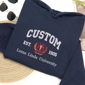 Könnte beinhalten: Marineblauer Kapuzenpullover mit dem Wort "CUSTOM" in Weiß gestickt. Darunter steht "EST. 1905" mit einem Wappen und "Loma Linda University" in Weiß. Der Pullover hat eine Kapuze mit Kordelzug.