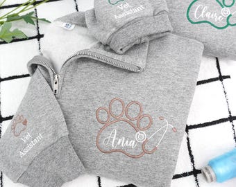 Könnte beinhalten: Graues Sweatshirt mit Viertelreißverschluss und gestickten Pfotenabdrücken und Text. Ein Pfotenabdruck ist in Braun mit dem Namen "Ania" und einem Stethoskop gestickt. Ein weiterer Pfotenabdruck ist grün mit dem Namen "Claire". Die Ärmel sind mit "Vet Assistant" bestickt.