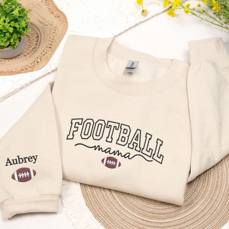 Custom embroidered Football Mama sweatshirt in Sand, featuring varsity-style lettering with football design and personalized sleeve name 'Aubrey'. Cozy cotton-poly blend for comfort and durability. Perfect gift for football moms, game day outfits, or showing team spirit. Handmade embroidery creates a premium, one-of-a-kind keepsake. Available in multiple colors and sizes