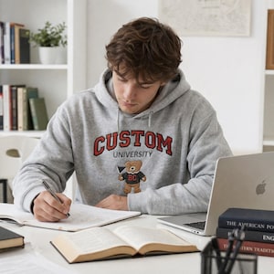A young man studying at a desk wearing a Gildan Sport Grey heavy blend hoodie. The chest features high-quality custom university embroidery with "CUSTOM" in red block letters and a cute embroidered teddy bear mascot. This premium personalized collegiate hoodie is perfect for student life, study sessions, and campus fashion enthusiasts looking for unique, high-density stitching.