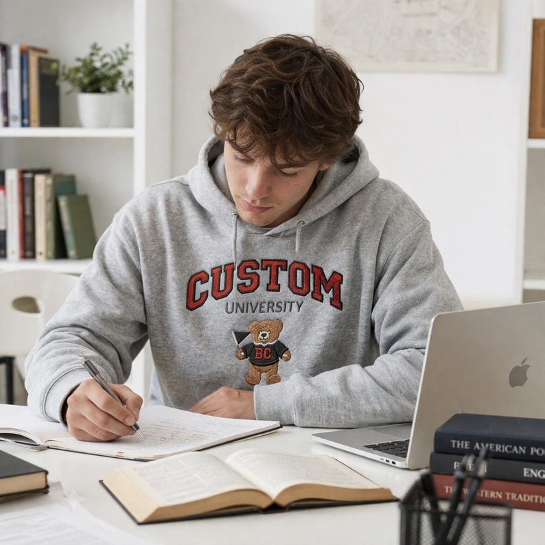 A young man studying at a desk wearing a Gildan Sport Grey heavy blend hoodie. The chest features high-quality custom university embroidery with "CUSTOM" in red block letters and a cute embroidered teddy bear mascot. This premium personalized collegiate hoodie is perfect for student life, study sessions, and campus fashion enthusiasts looking for unique, high-density stitching.