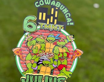May include: A colorful birthday cake topper featuring the Teenage Mutant Ninja Turtles. The design includes the number 6, the word "COWABUNGA!", and the name "JULIUS". The turtles are holding pizza.