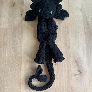 Può includere: Peluche di drago nero all'uncinetto con occhi verdi. Il drago è sdraiato sulla schiena con le gambe e la coda arrotolate.