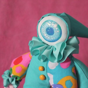 May include: A whimsical handmade doll with a large, detailed eye. The doll features a teal outfit with a ruffled collar and a pointed hat. The arms have colorful, patterned fabric. Two orange buttons adorn the front.
