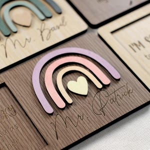 May include: A wooden plaque with a rainbow design and the text "Mr. Patrick" engraved on it. The rainbow is made of pastel pink, peach, and lavender colors. A small heart is in the center of the rainbow.