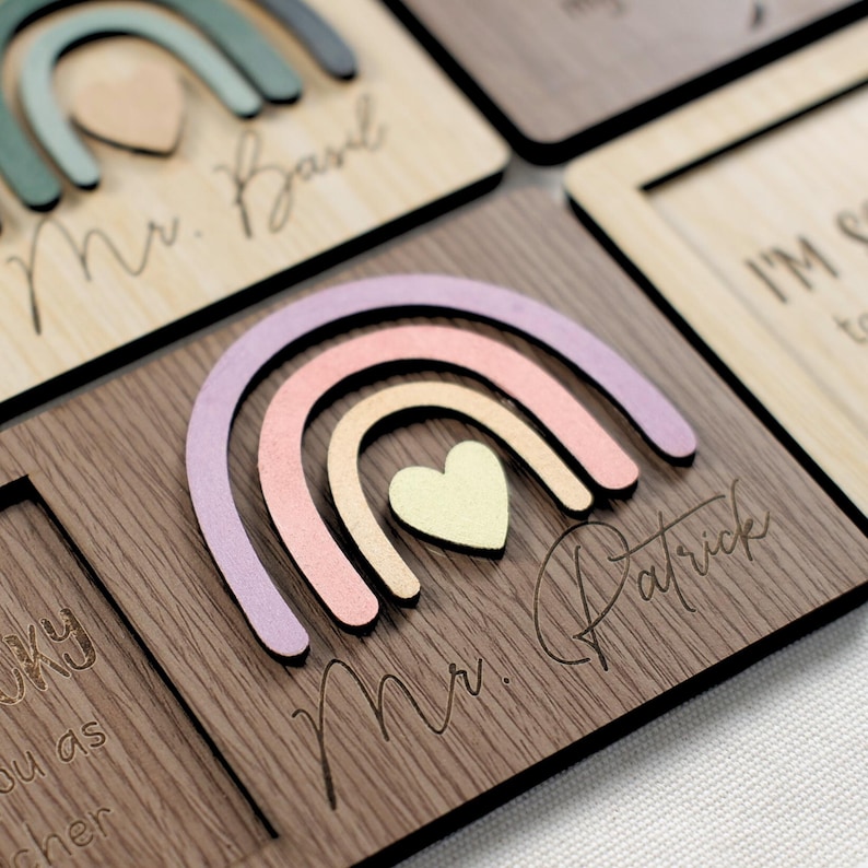 May include: A wooden plaque with a rainbow design and the text "Mr. Patrick" engraved on it. The rainbow is made of pastel pink, peach, and lavender colors. A small heart is in the center of the rainbow.