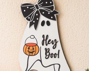 May include: A white wooden ghost-shaped Halloween decoration with a black bow and the words "Hey Boo!" The ghost holds an orange pumpkin bucket. The bow has white polka dots. The decoration is hanging from a white string.