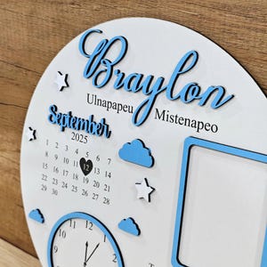 May include: A white, circular wooden plaque with blue accents. The plaque features the name "Braylon" in cursive, the month "September" and the year "2025". It includes a calendar, clock, clouds, stars, and a photo frame.