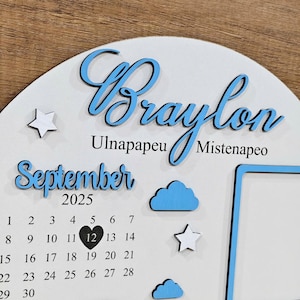 May include: A white wooden baby milestone calendar with the name "Braylon" in blue script. The calendar includes the month "September 2025", a calendar grid, and decorative stars and clouds. There is a photo frame on the right.