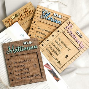 May include: Four wooden picture frames with a quote about teaching. The frames are decorated with a wood grain pattern and have a black and white checkered border. The quote reads "The wonder of teaching is watching caterpillars become butterflies". Each frame has a different teacher's name on it: Ms. Mattianna, Ms. Parabella, Ms. Florian, and Ms. Rosemary.