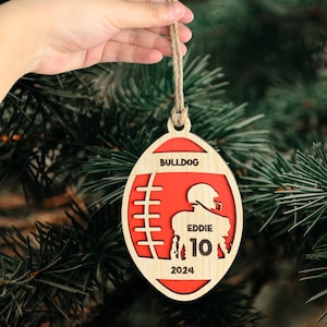 May include: A wooden football ornament with a red background and the text "BULLDOG" at the top. The ornament also features a silhouette of a football player with the name "EDDIE" and the number "10" on the jersey. The year "2024" is printed at the bottom of the ornament.