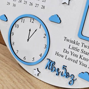 May include: A white wooden clock with a blue border and hands pointing to 1:05. The clock has blue cloud and star accents. Text includes "Twinkle Twinkle Little Star" and "Mrs. Doz".