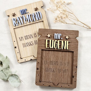 Op de afbeelding: Twee houten borden in de vorm van een pot met een stervormig ontwerp. Het bovenste bord heeft de tekst "Mr. Raymond" en het onderste bord heeft de tekst "Mr. Eugene". Beide borden hebben de tekst "My brain is full thanks to you".