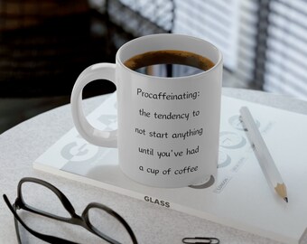 May include: White ceramic coffee mug with black text that reads "Procaffeinating: the tendency to not start anything until you've had a cup of coffee". The mug is filled with coffee and is sitting on a white surface with a notebook, a pencil, and glasses.
